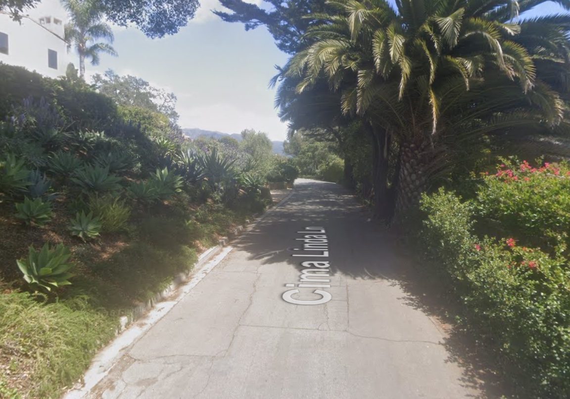 Cima Linda Lane: a narrow paved lane with a low curb, planted slope and palms. Google Street View, April 2025.